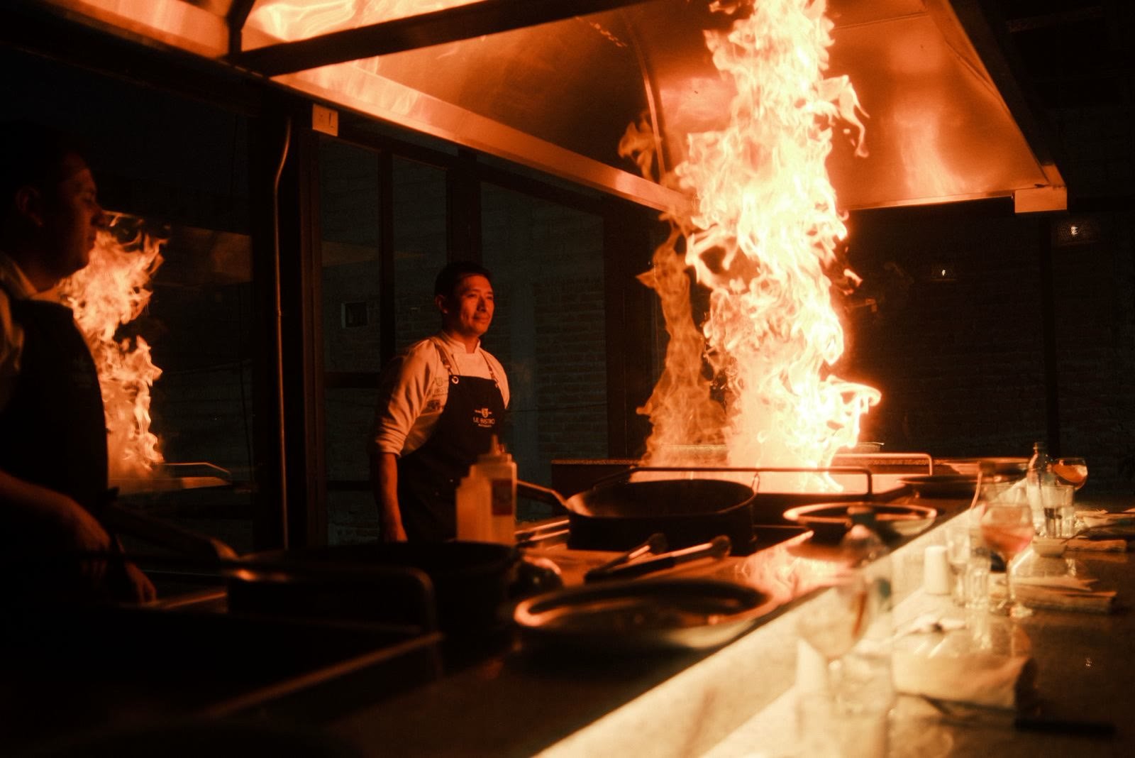 Llamarada alta sobre la plancha frente a la barra, con un chef observando